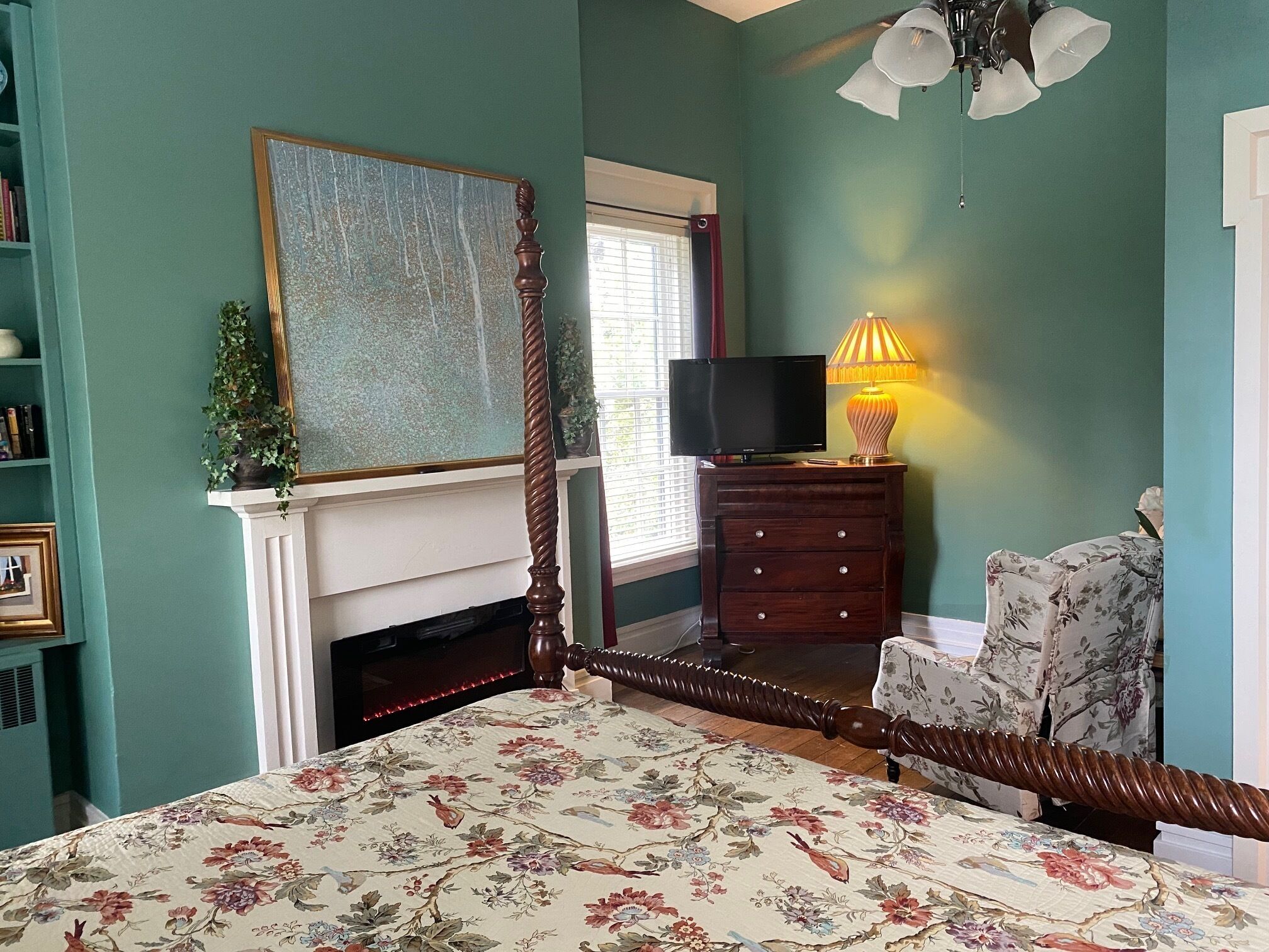 Quarto com cama de dossel, televisão sobre uma cômoda, lareira e janela com persianas.