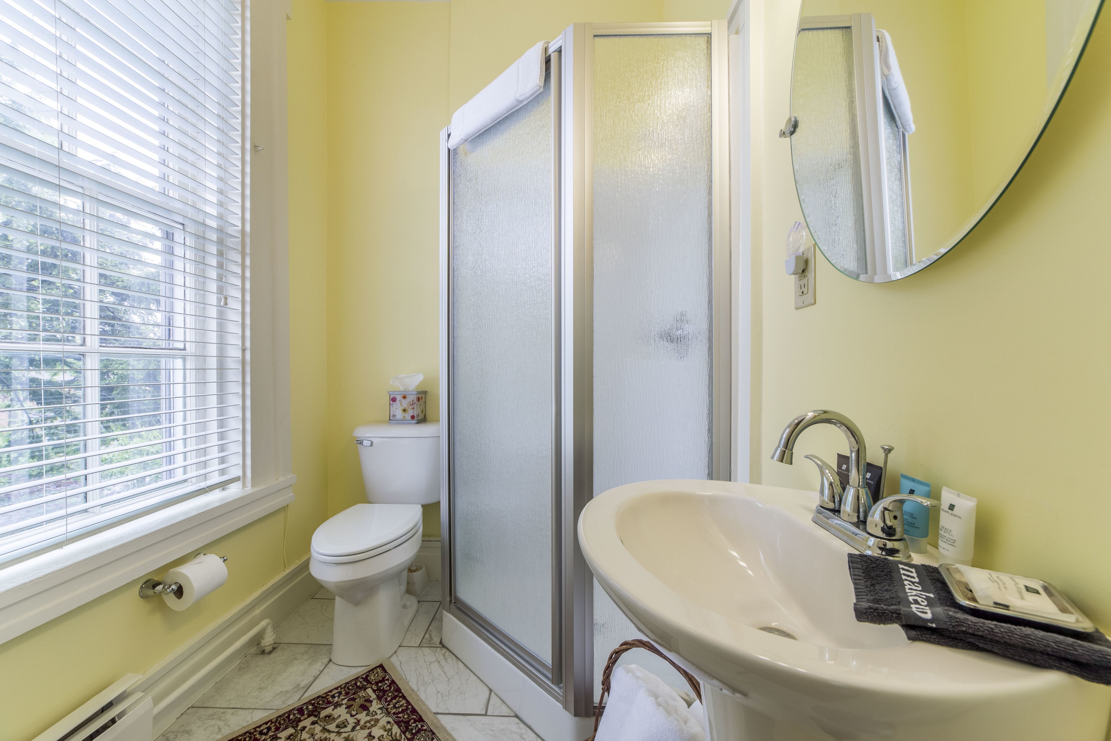 A bathroom with a toilet, a shower enclosure, a sink, and a window with blinds.