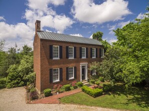 A two-story brick house with a metal roof, a front door, and a small front yard with a pathway and some shrubs.