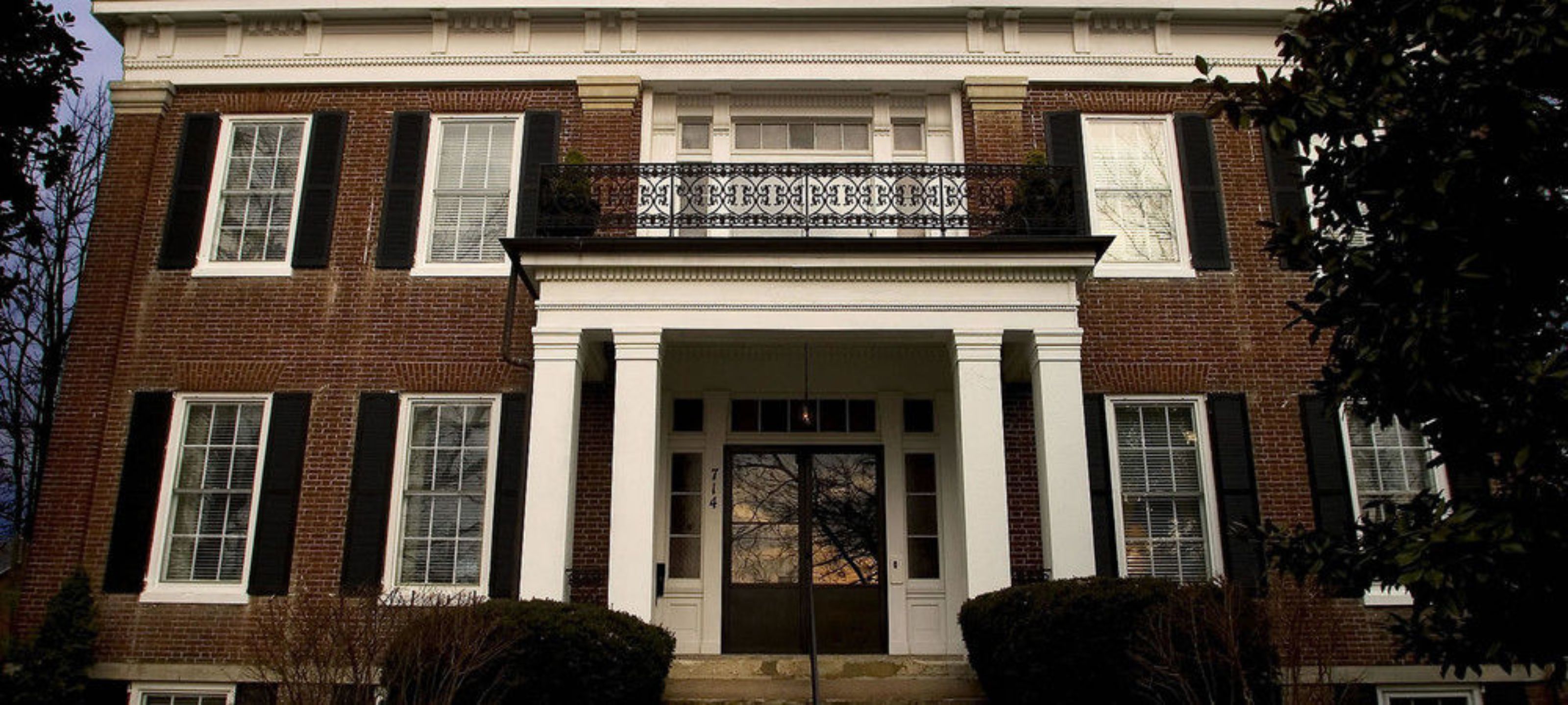 A two-story brick building with a central entrance flanked by columns, a balcony with decorative railings, and a number plaque.