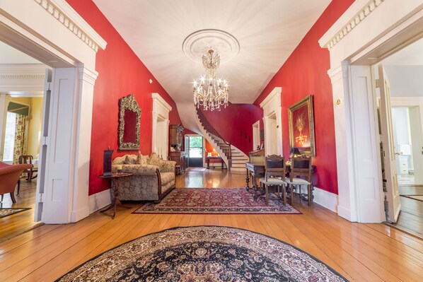 A spacious living room with a red accent wall, a chandelier, a staircase, and a dining area. - Bourbon Manor B&B (Bardstown)