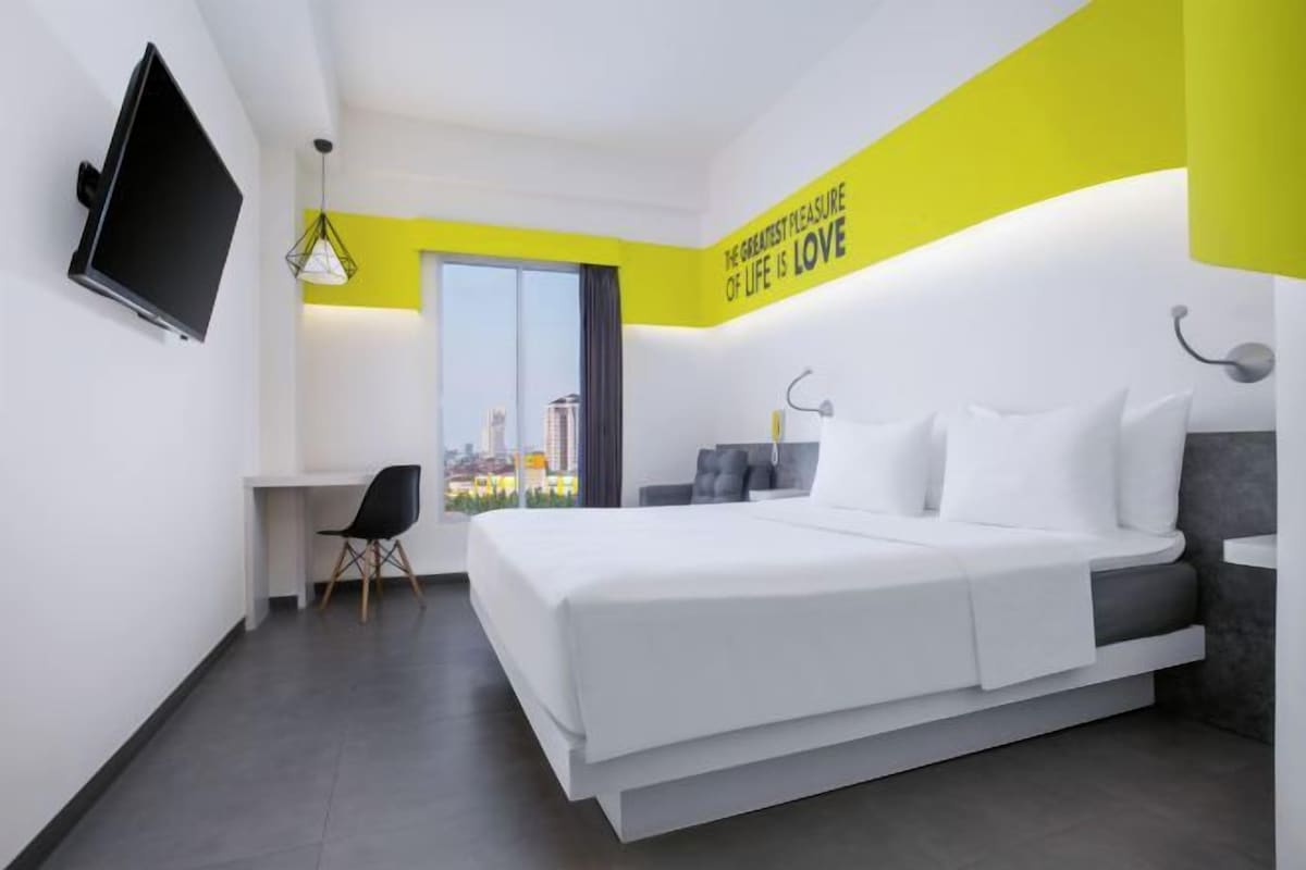Una habitación de hotel moderna con cama, escritorio, silla y un amplio ventanal con vistas a la ciudad.