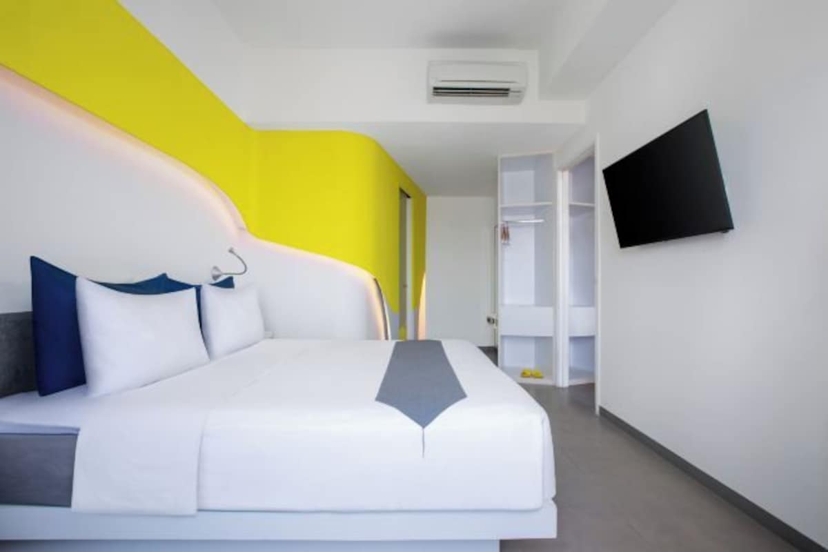 A modern hotel room with a white bed, a bright yellow accent wall, and a flat-screen TV mounted on the wall.