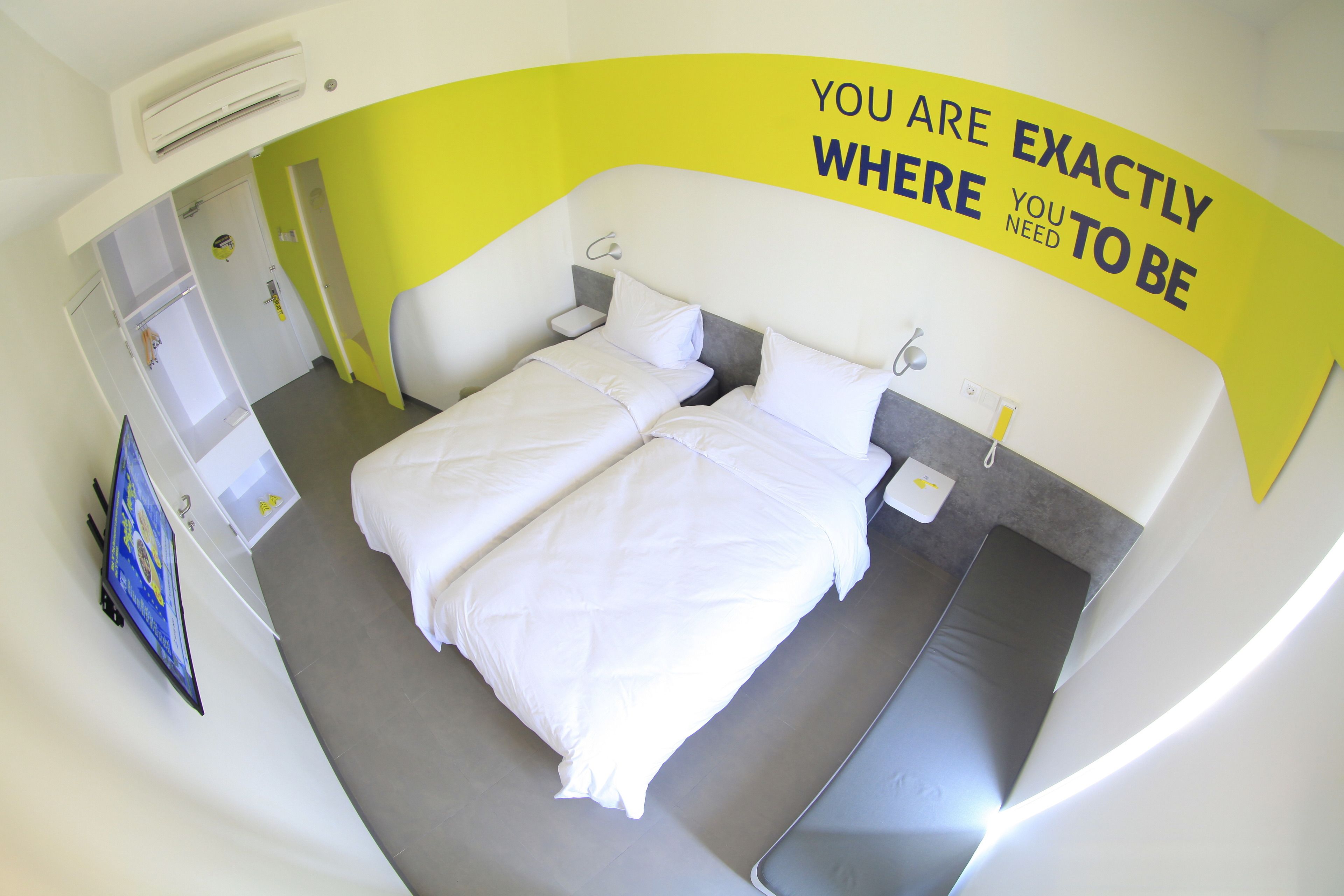 A hotel room with a bed, bedside tables, an air conditioning unit, and a motivational wall message.