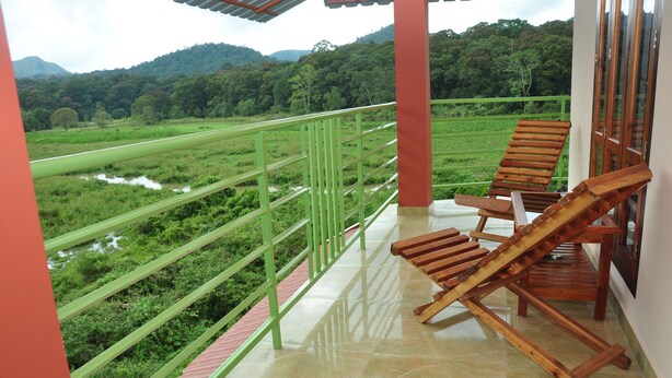A balcony with a view of a lush green landscape and a river. There are two wooden chairs and a table on the balcony. - Kerala House (Peermade)