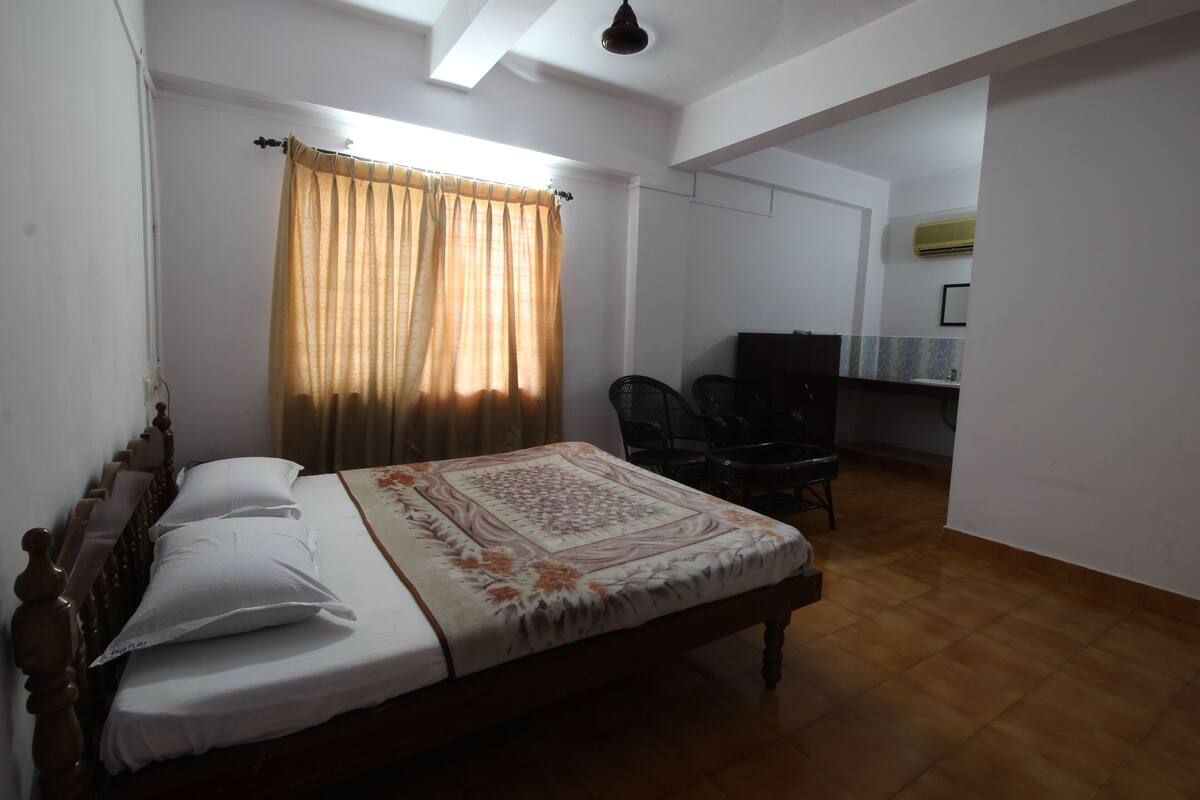 A bedroom with a wooden bed, a window with curtains, a desk with a chair, and a bathroom with a mirror and sink.