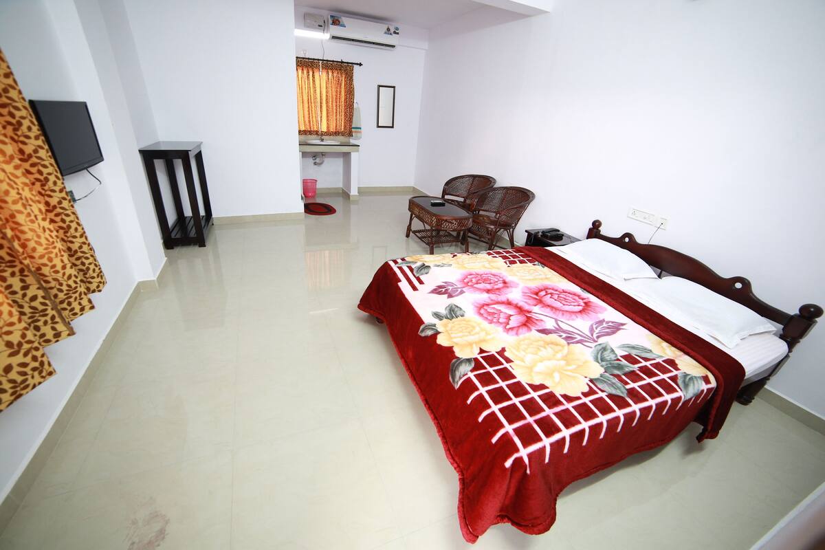 A hotel room with a bed, a small table, a chair, a television, and a bathroom visible in the background.