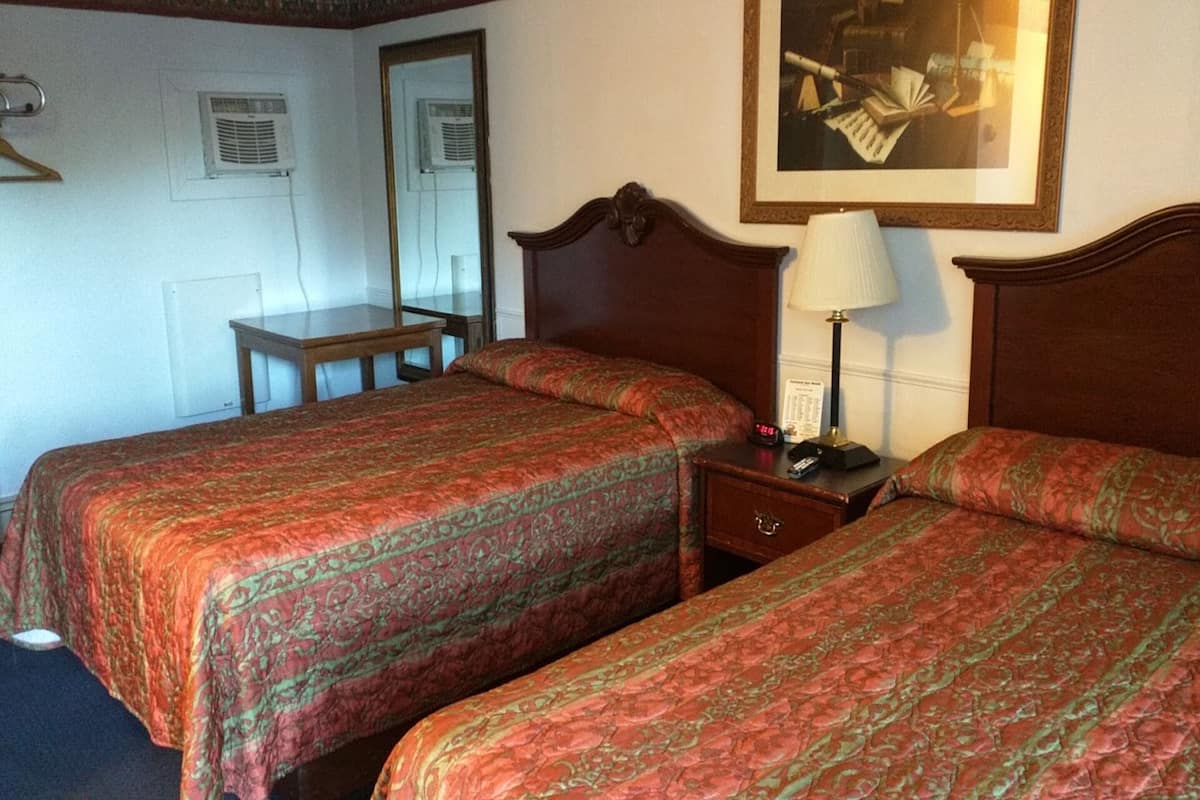 A hotel room with two beds, a bedside table with a lamp, a chair, and a painting on the wall.