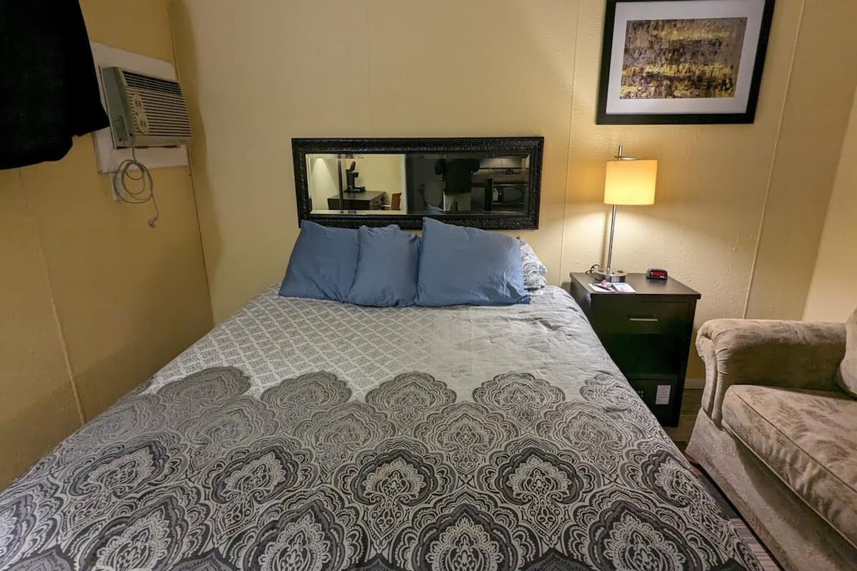 A hotel room with a bed, a nightstand, a lamp, a chair, and a wall-mounted air conditioner.