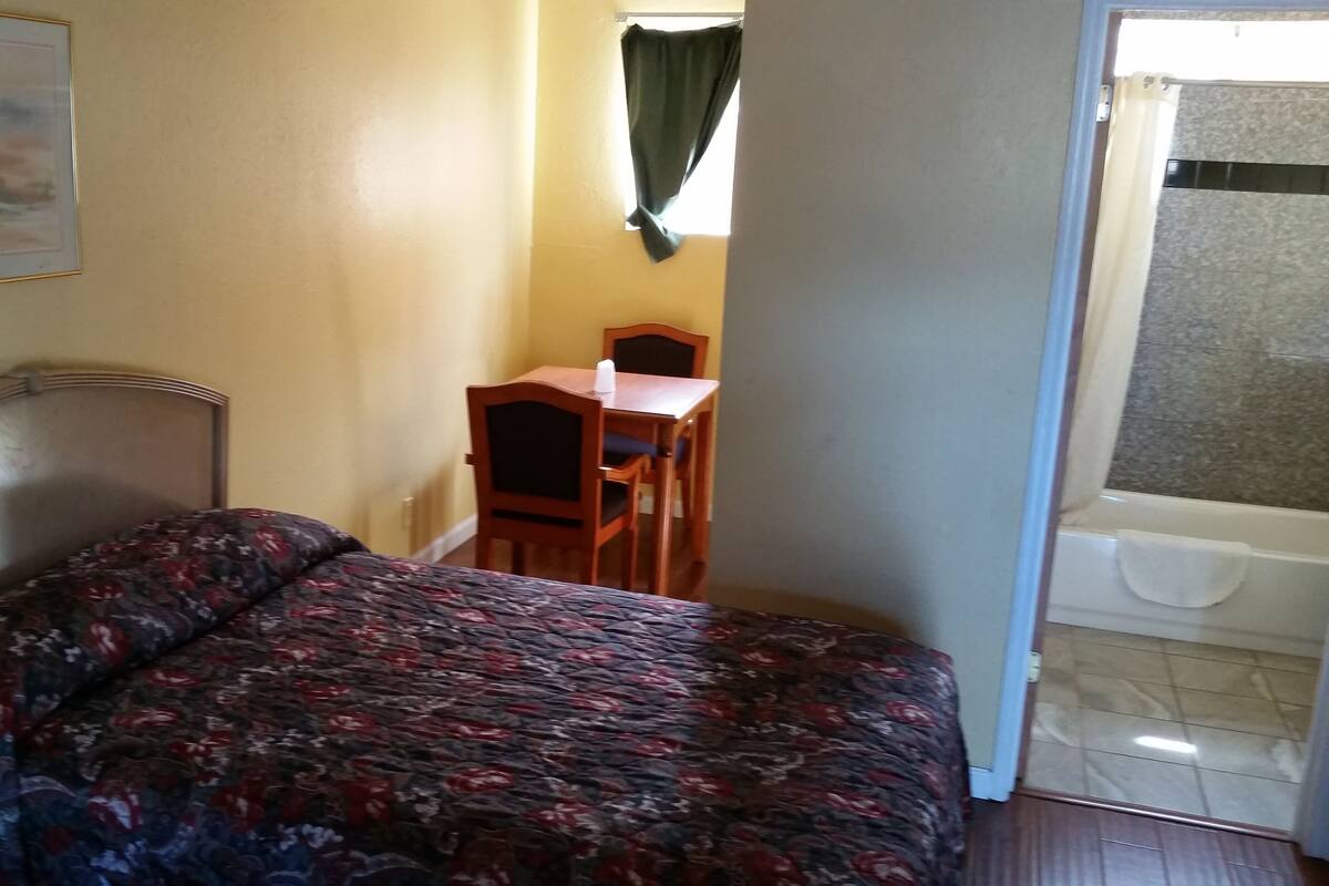 A small, compact room with a bed, a desk, and a chair. There is a window with a curtain, and a bathtub is visible through the door.