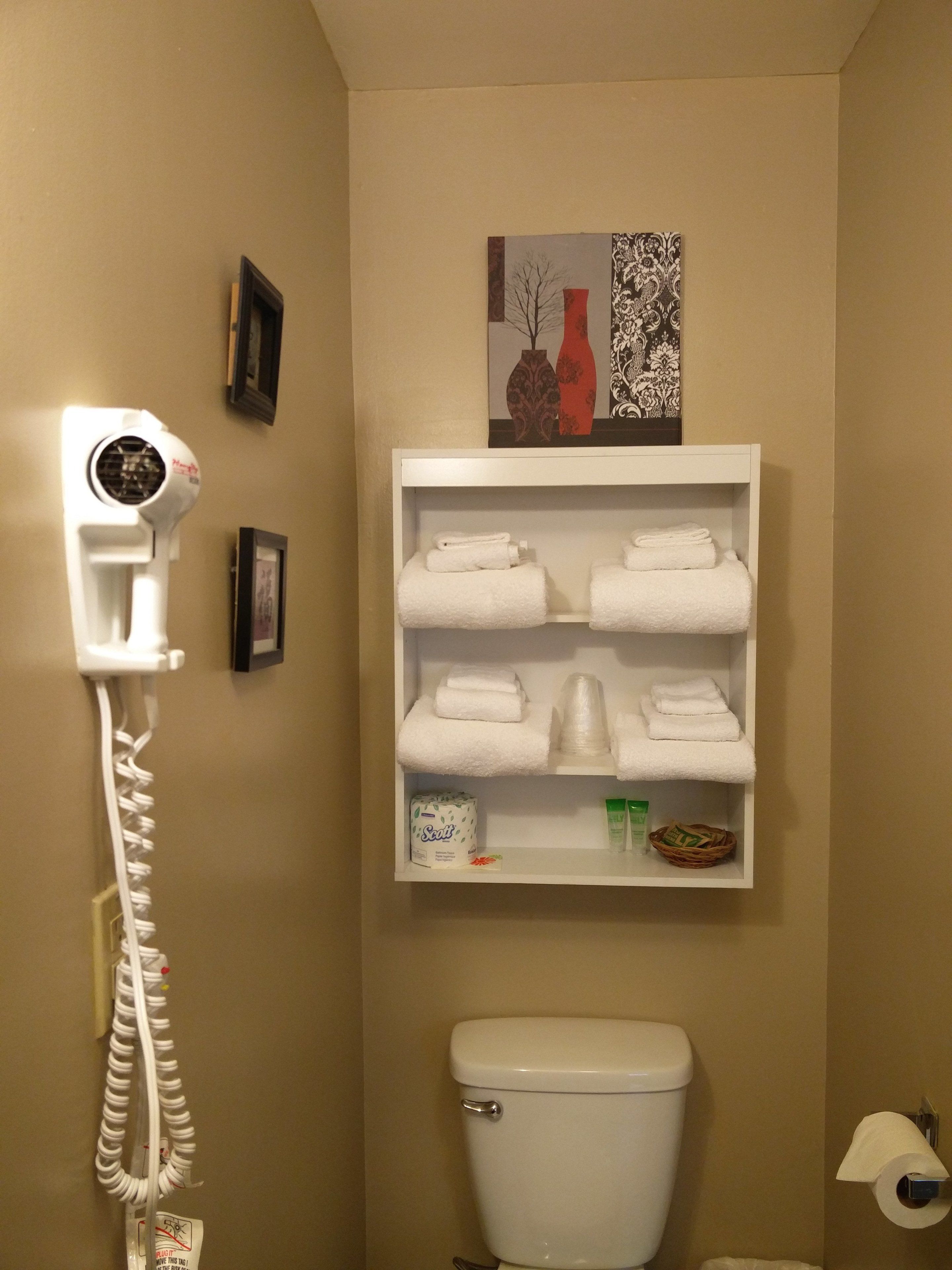 A bathroom with a toilet, a shelf with folded towels, a hairdryer, and a toilet paper holder.