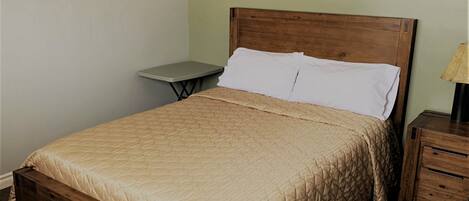 Una cama individual con estructura de madera y una colcha beige acolchada. Hay una mesita de noche pequeña con una lámpara encima. La pared está pintada de verde claro, y hay un cuadro enmarcado sobre la cama.