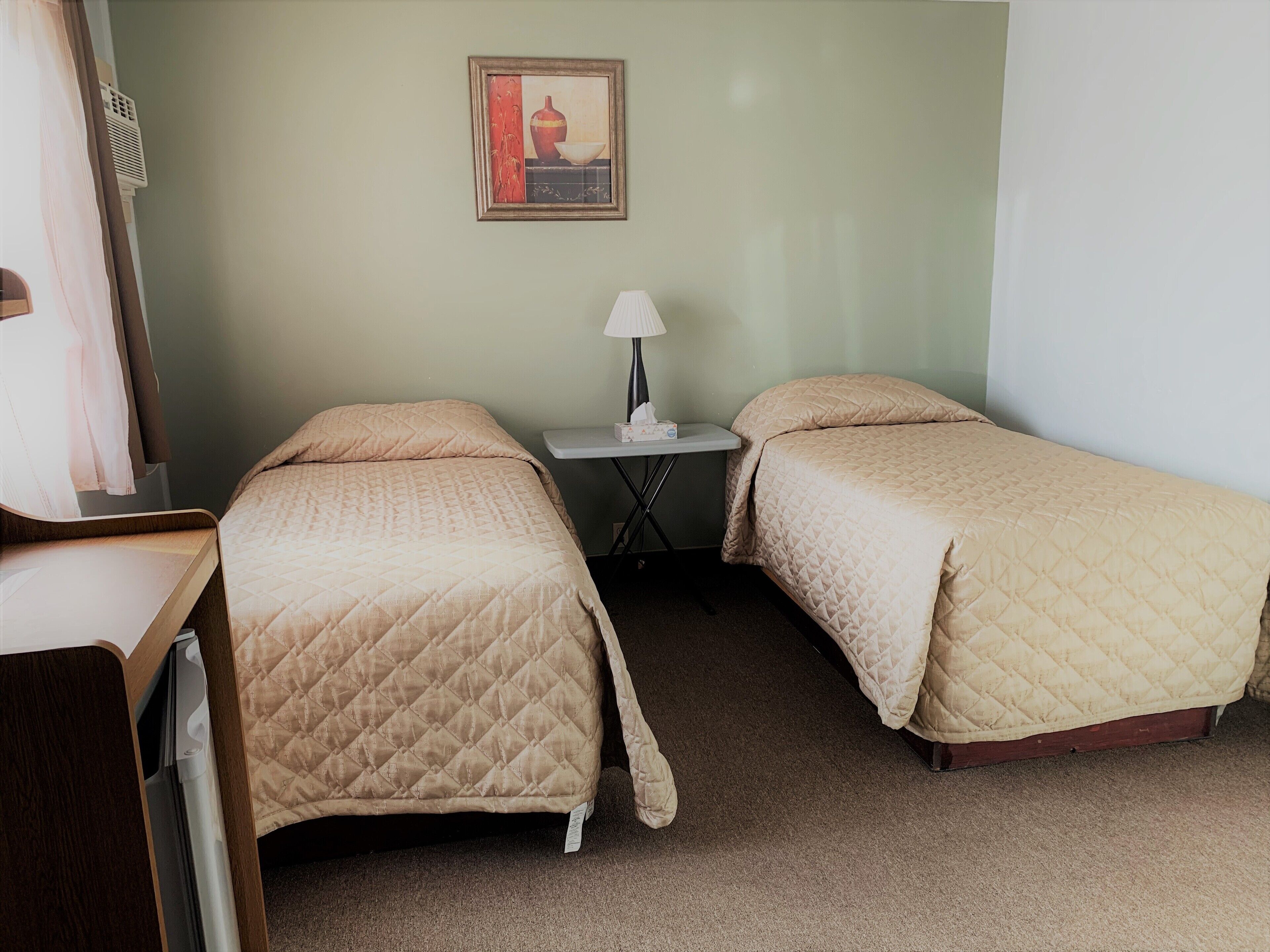 A small, neatly arranged room with two single beds, a small table with a lamp, and a wall-mounted air conditioning unit.