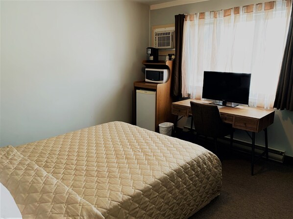 Una habitación de hotel con cama, escritorio con computadora, refrigerador pequeño y aire acondicionado.
