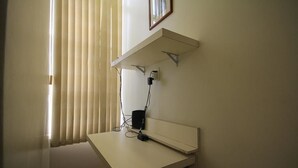 A room with a white desk, a wall-mounted shelf, and a window with blinds.