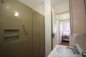 A bathroom with a glass shower enclosure, a sink with a mirror, and a bed visible through an open door. - Domingos 1101 (Rio de Janeiro)