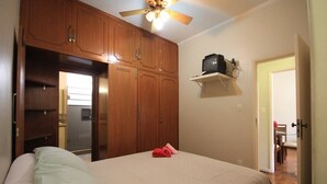 A bedroom with a bed, wooden wardrobe, ceiling fan, and a small shelf with a TV.