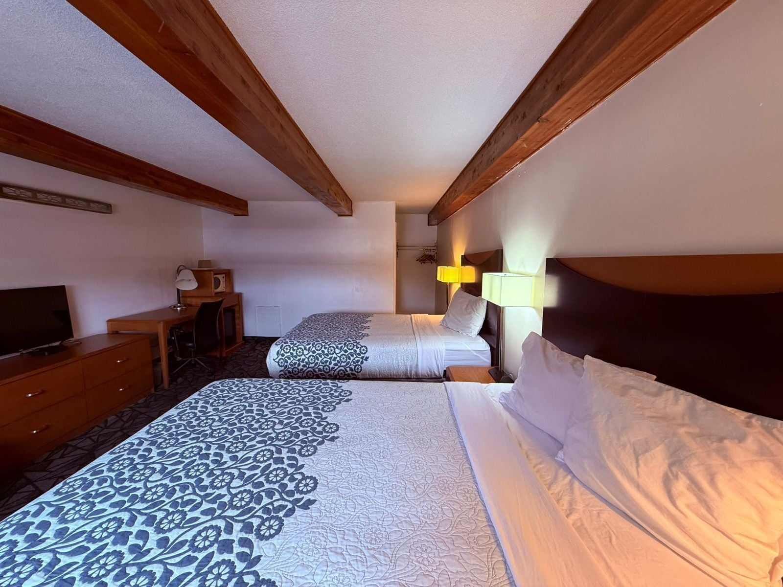A hotel room with two beds, wooden beams on the ceiling, a desk with a lamp, and a television.