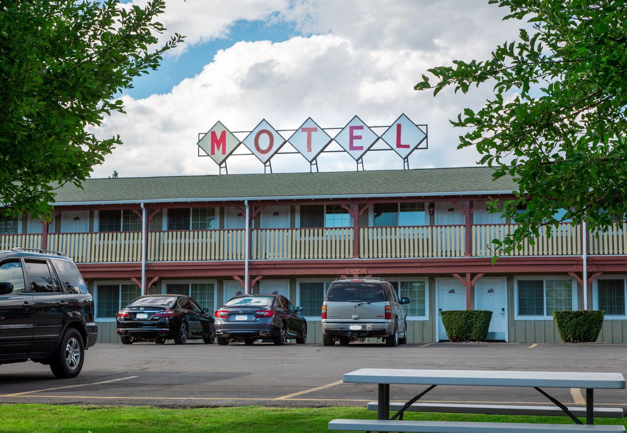A motel with a row of parked cars in front, a picnic table, and a signboard spelling out "MOTEL".