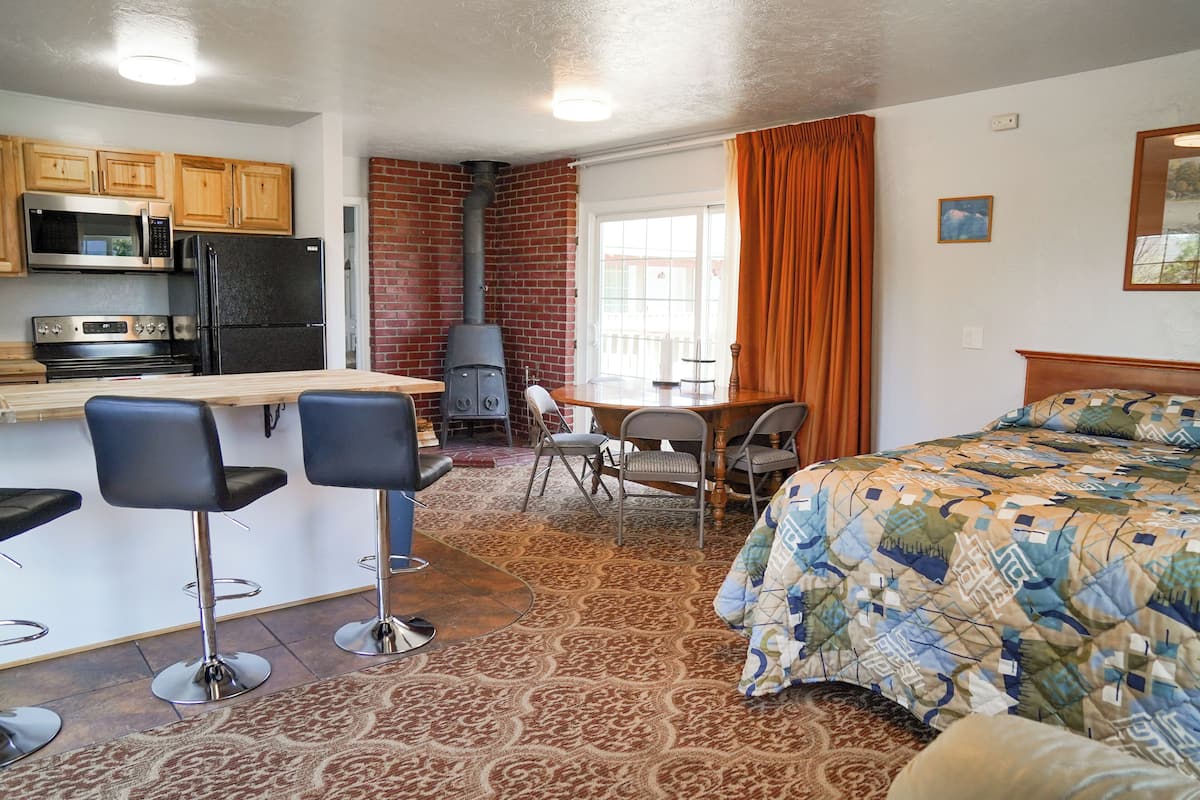 A compact kitchen with a microwave, stove, and refrigerator. There is a dining area with a table and chairs, and a bed with a patterned blanket.