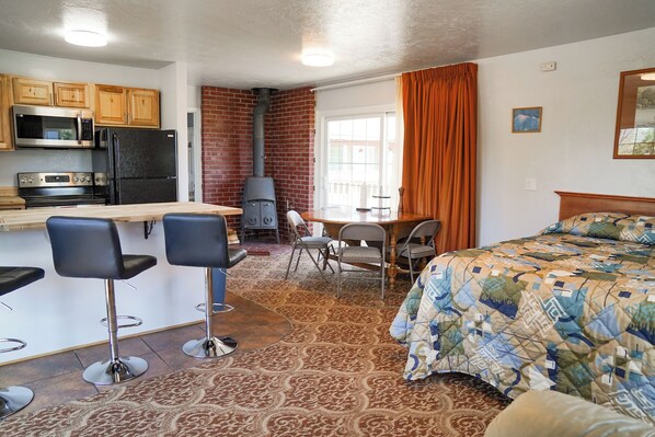 A compact kitchen with a microwave, stove, and refrigerator. There is a dining area with a table and chairs, and a bed with a patterned blanket. - Swiss Holiday Lodge (Mount Shasta)
