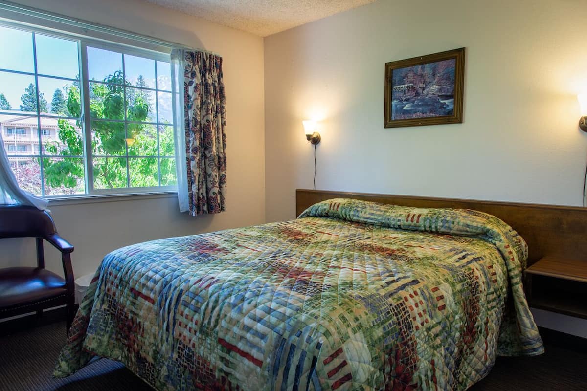 Quarto de hotel com uma cama grande, uma cadeira, janela com cortinas, um quadro na parede e duas luminárias de parede.