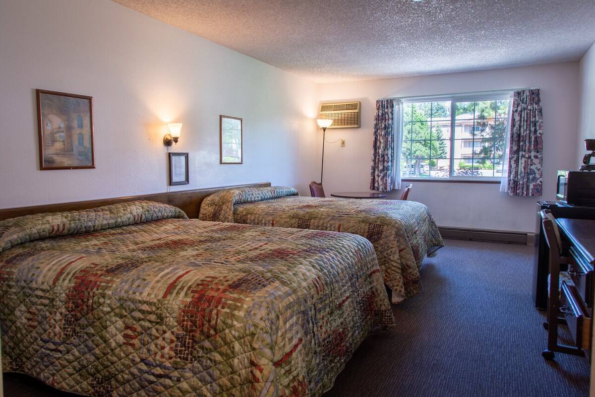 Quarto de hotel com duas camas, uma escrivaninha, uma luminária e uma janela com cortinas.