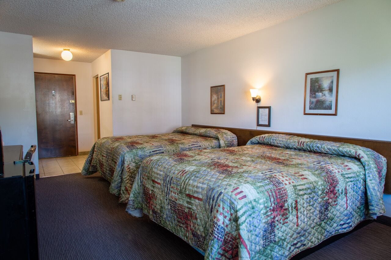 A hotel room with two beds, a door, a wall-mounted light, and framed pictures.