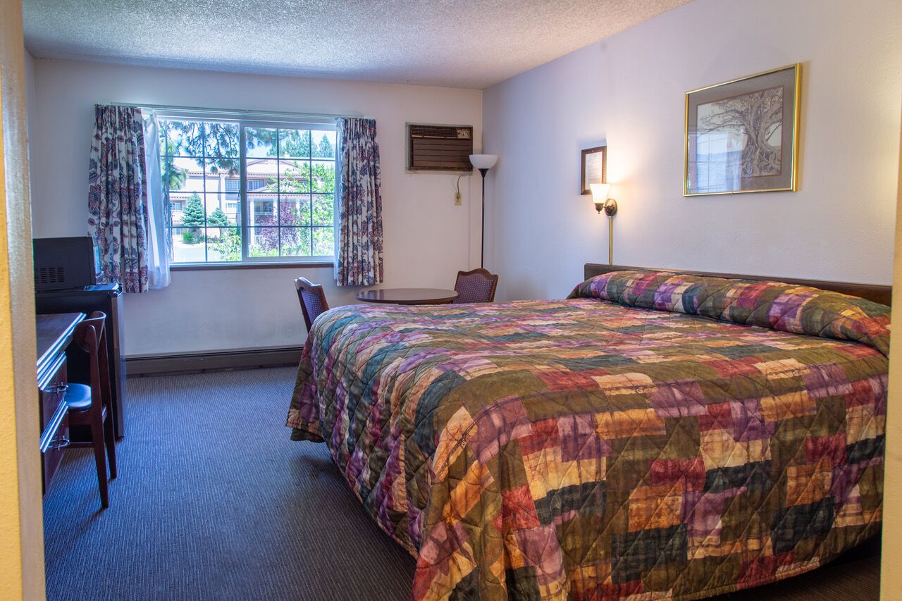 A hotel room with a large bed, a desk with a chair, a window with curtains, a floor lamp, and a framed picture on the wall.