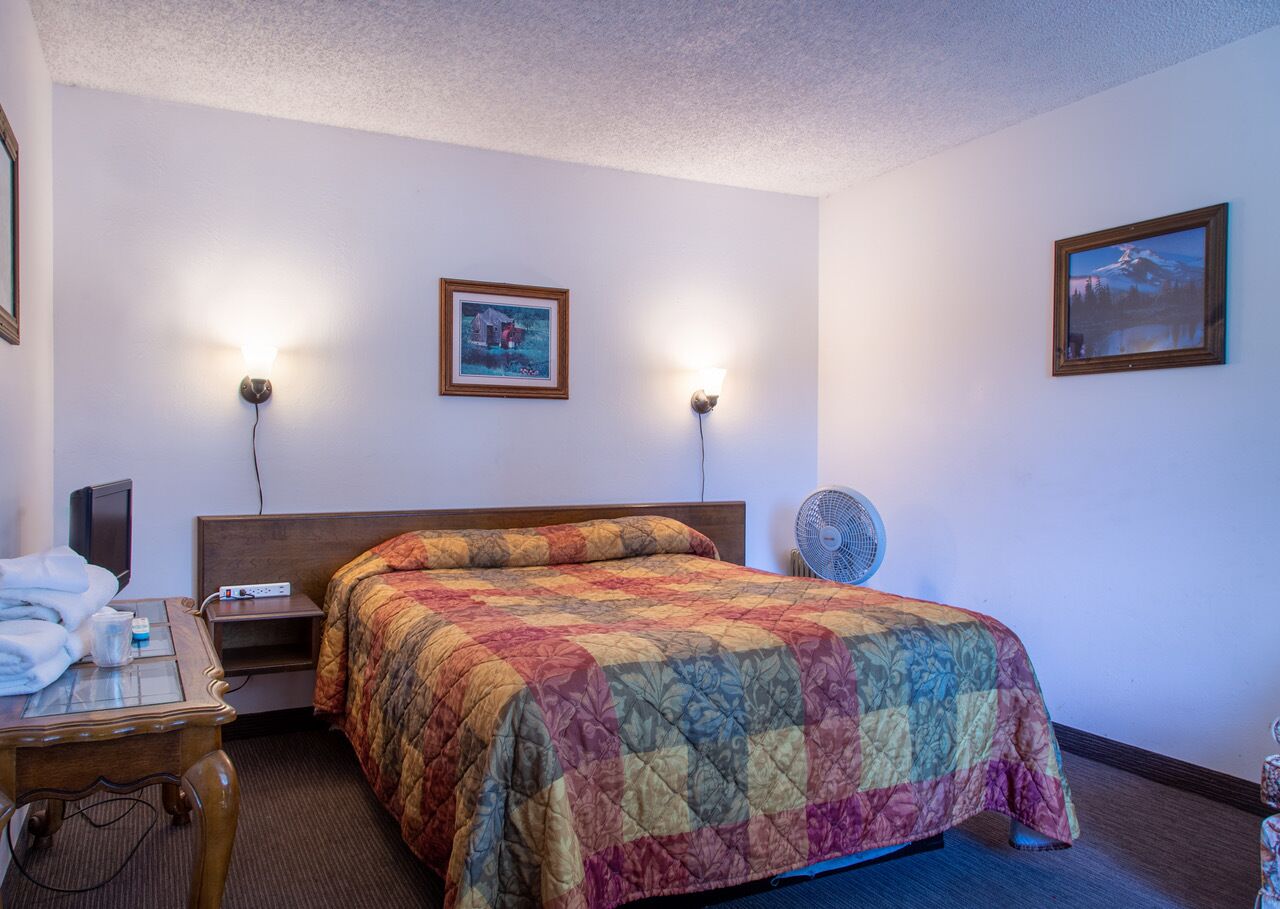 A hotel room with a large bed, two wall-mounted lamps, a bedside table with a lamp, a small table with towels, and a wall-mounted TV.