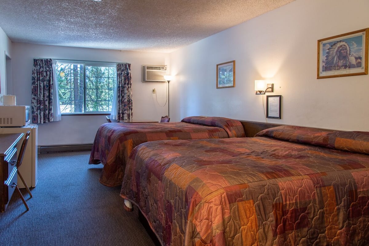 Quarto de hotel com duas camas, uma escrivaninha, uma cadeira, janela com cortinas e luminárias na parede.