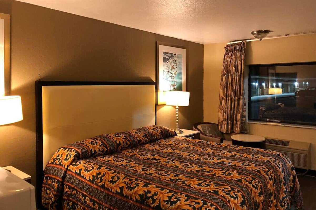 Une chambre d’hôtel avec un lit, un bureau doté d’une lampe, une chaise et une grande fenêtre avec des rideaux.