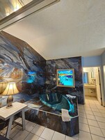Un baño con pared de mármol, una bañera, un televisor y una mesa con lámpara.
