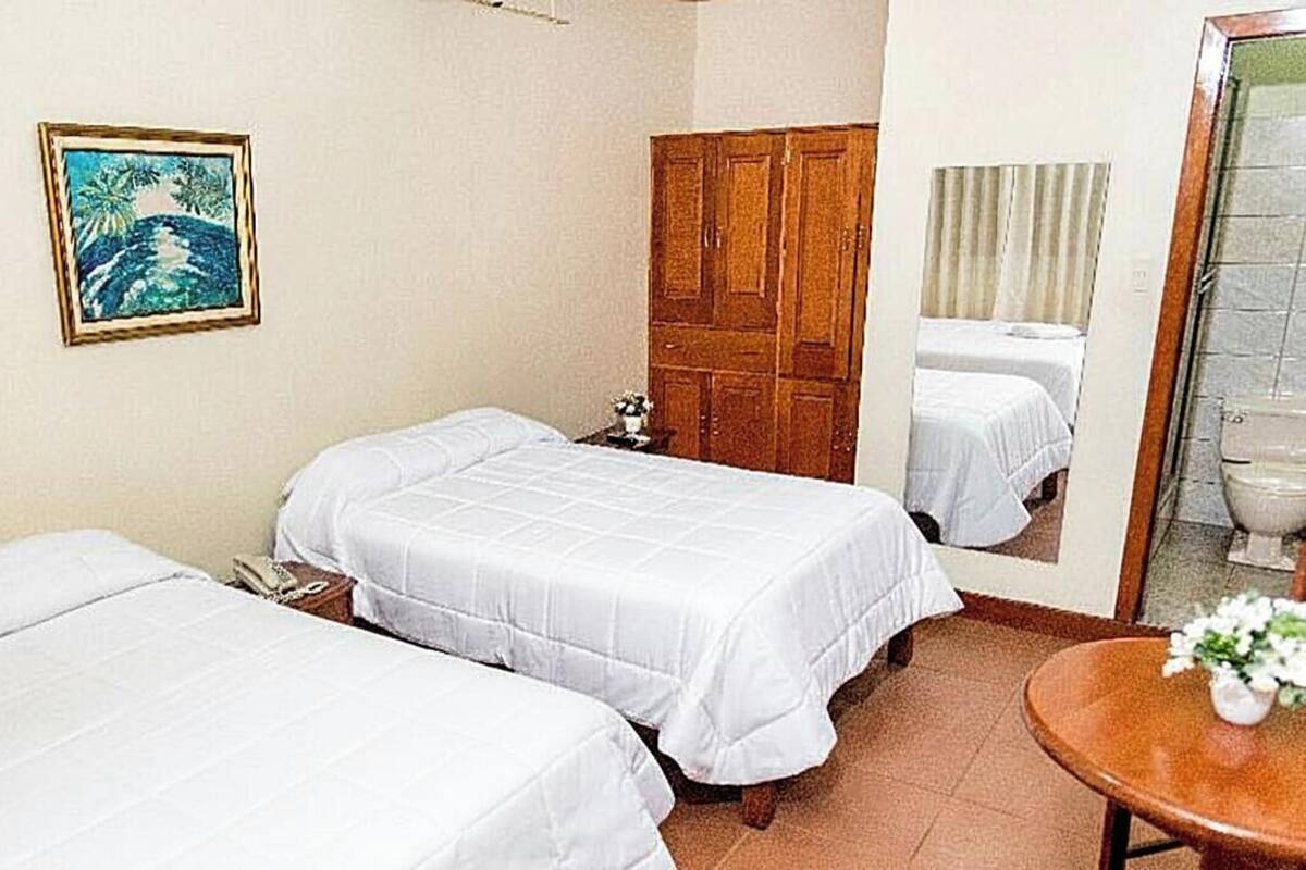 A hotel room with two beds, a wooden wardrobe, a painting on the wall, and a bathroom visible through a glass door.