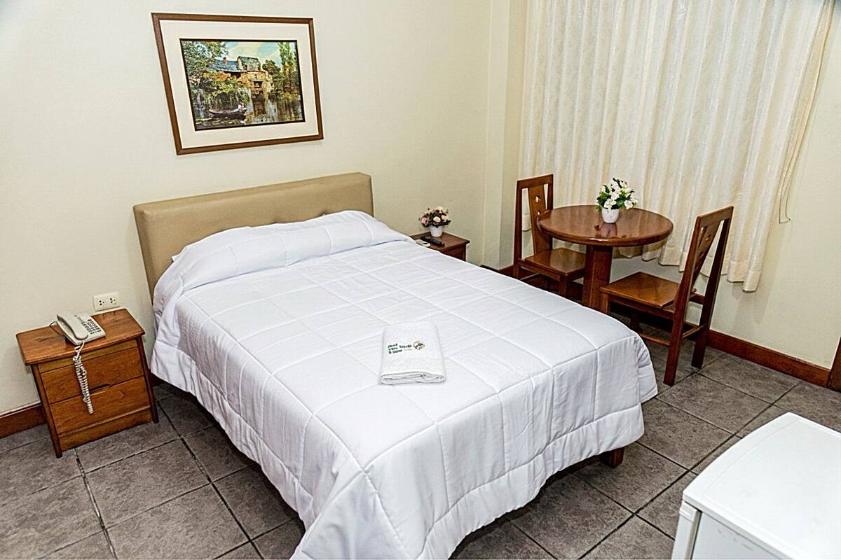 A neatly made bed with a white comforter and a folded towel on top. There is a small bedside table with a phone and a vase of flowers. A round wooden table with two chairs is positioned near a window with curtains.