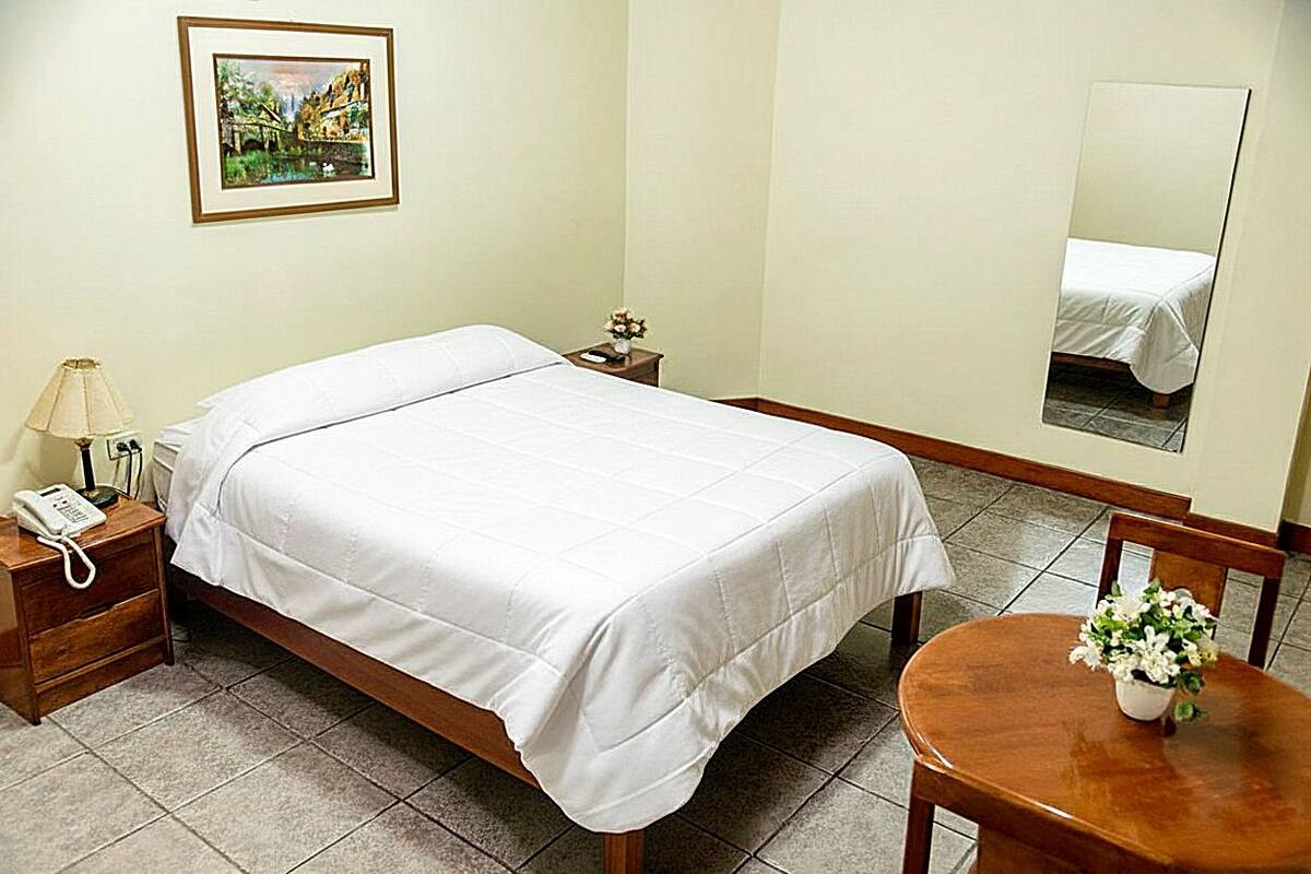 A hotel room with a bed, bedside table, lamp, and a small table with flowers.