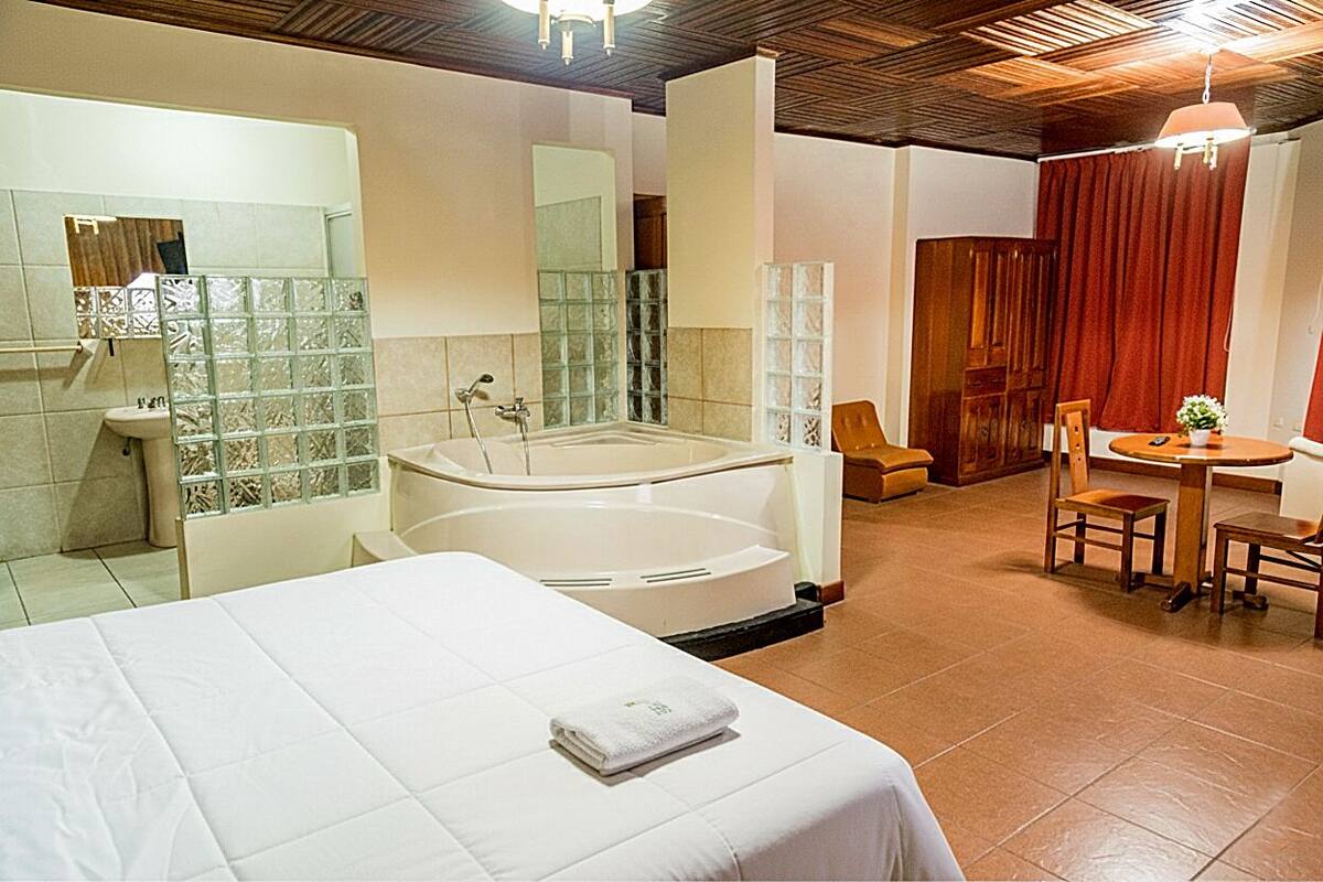 A hotel room with a large bed, a separate bathroom with a bathtub, a small dining area with a table and chairs, and a wooden ceiling.