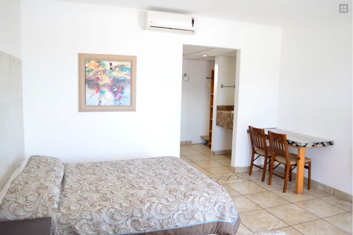 Una habitación de hotel con cama, escritorio y silla. Hay un cuadro en la pared y un equipo de aire acondicionado sobre la cama.