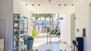 Un hall d’hôtel moderne avec une porte en verre donnant sur une cour intérieure agrémentée de palmiers et offrant une vue sur un bâtiment en arrière-plan.