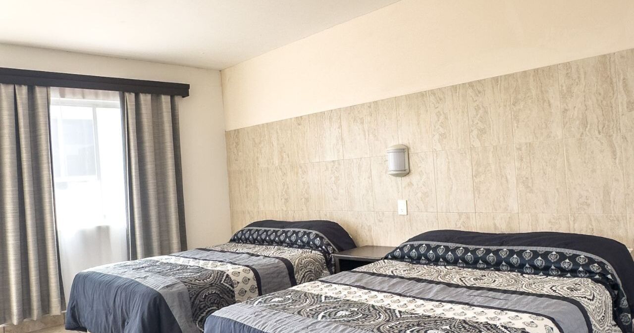 A hotel room with two beds, a large window with curtains, a wall-mounted light, and a tiled wall.