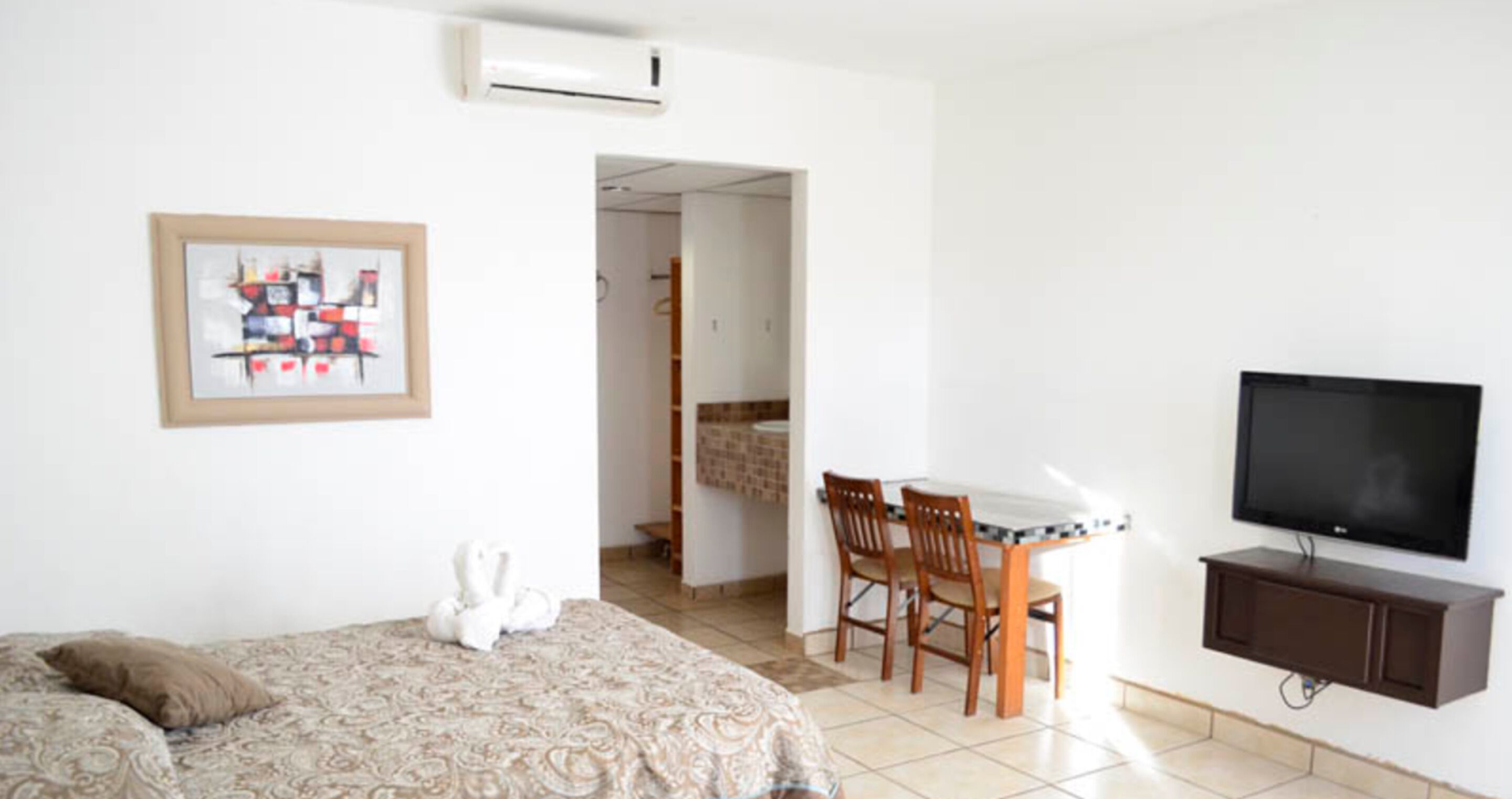A hotel room with a bed, a dining table with chairs, a television, and an air conditioning unit.