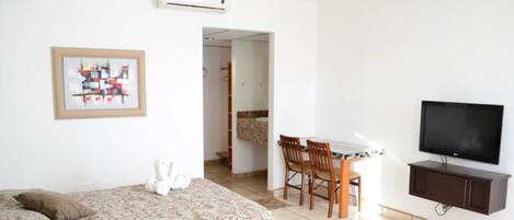 A hotel room with a bed, a dining table with chairs, a television, and an air conditioning unit.