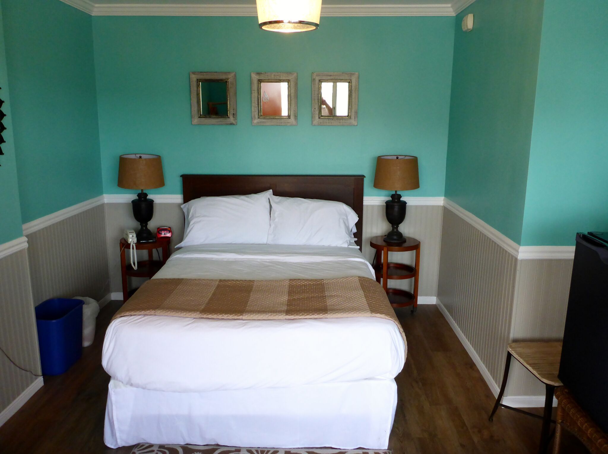 A neatly made bed in a room with teal walls, two bedside lamps, and three framed mirrors on the wall.