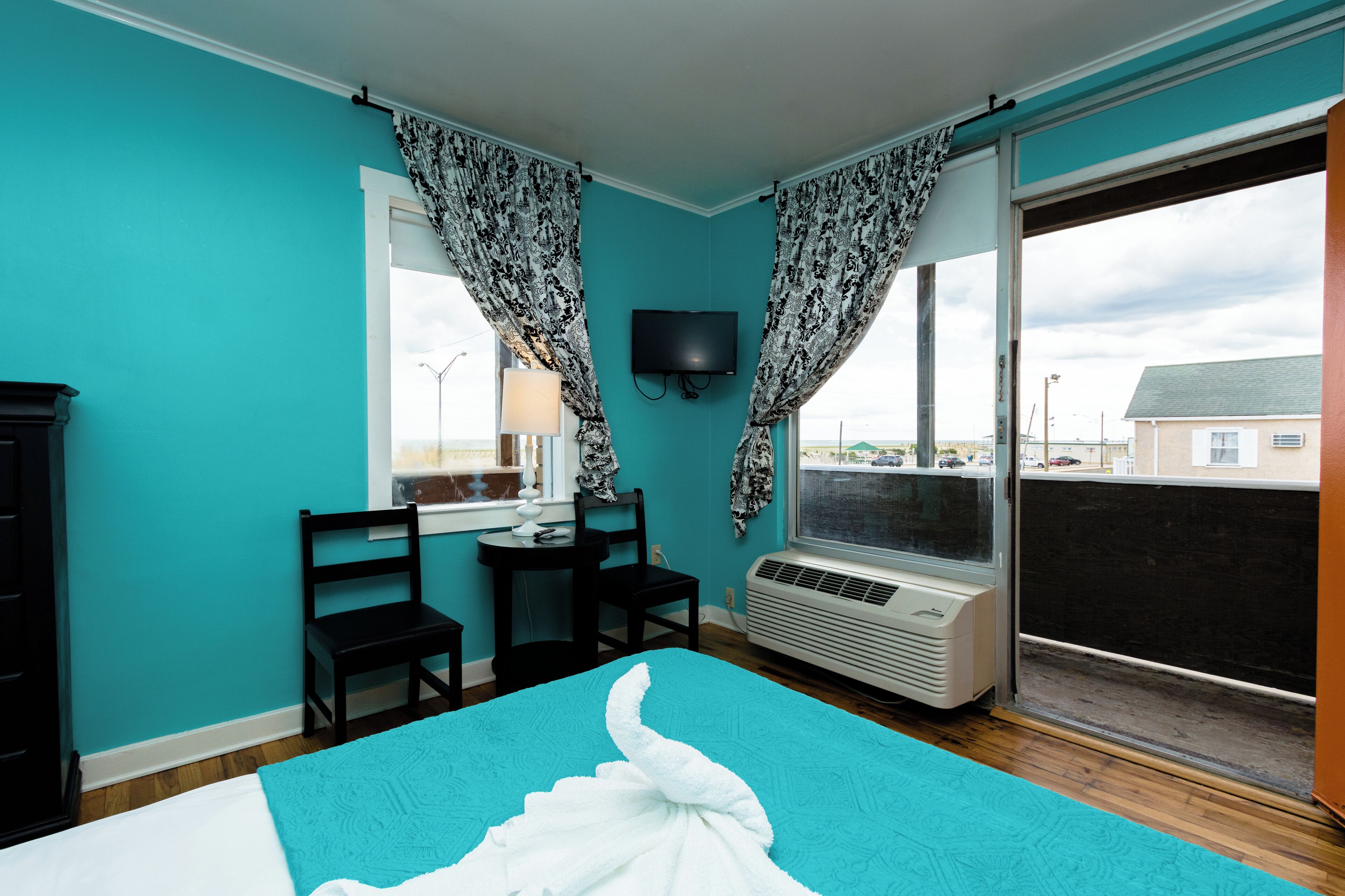A room with teal walls, a bed with a white and teal bedspread, two black chairs, a small table with a lamp, a TV mounted on the wall, and a large window with curtains.