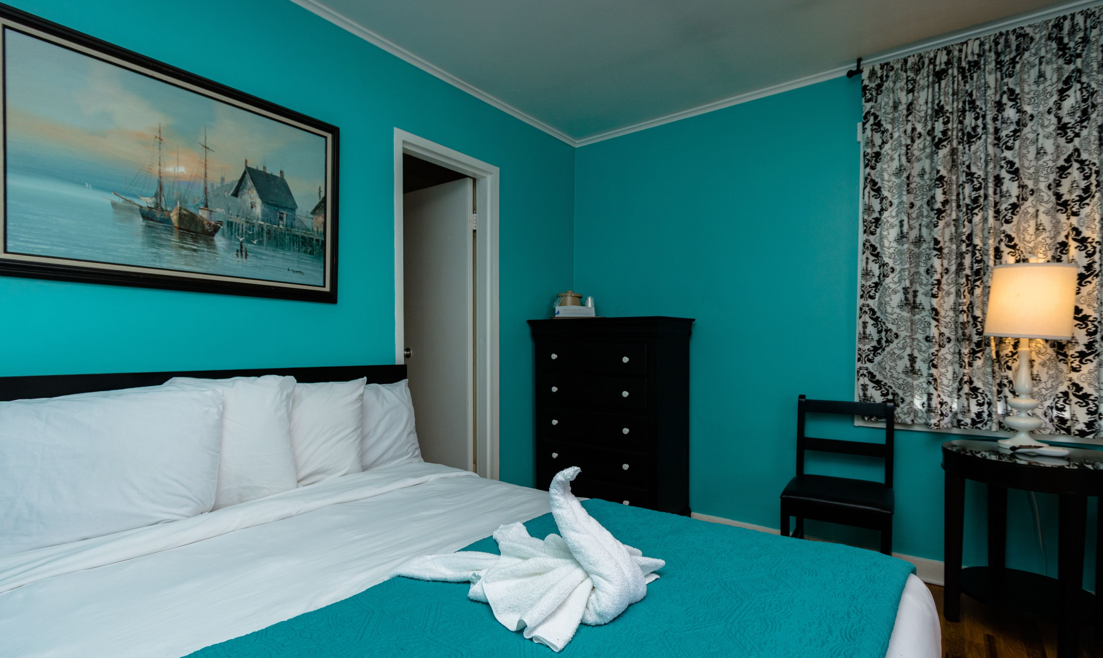 A hotel room with a bed, a painting of a coastal scene, a black dresser, a chair, and a lamp.