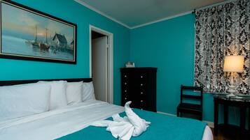 A hotel room with a bed, a painting of a coastal scene, a black dresser, a chair, and a lamp.