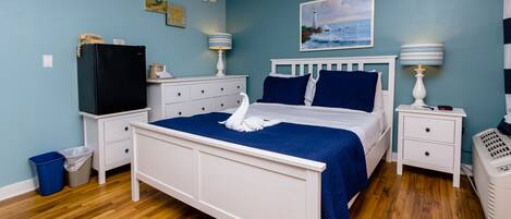 A bedroom with a bed, white nightstands, a heater, a trash bin, and a refrigerator.