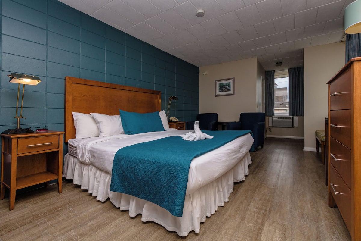A hotel room with a large bed, a wooden headboard, a nightstand with a lamp, a desk with a chair, and a wardrobe.