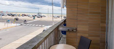 A balcony with wicker chairs and a small table, overlooking a beach with cars and a cyclist.