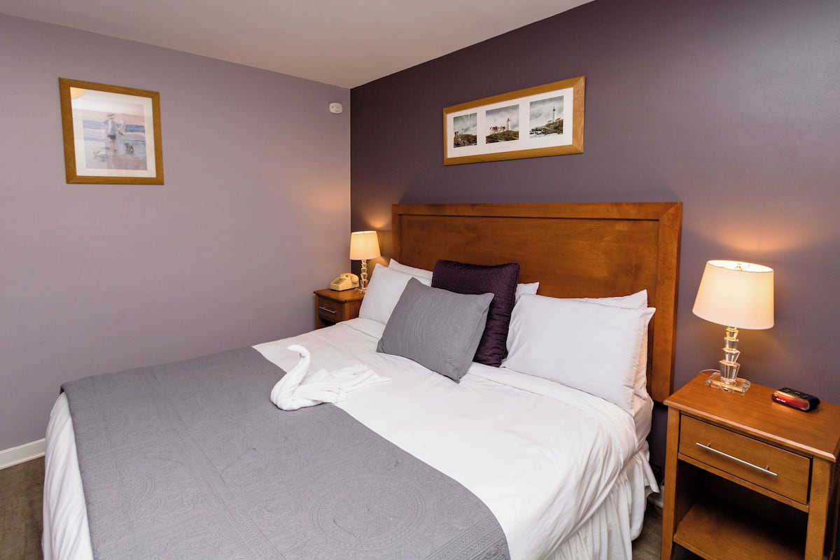 A neatly made bed with a white comforter and a gray throw blanket, two bedside lamps, a wooden nightstand with a phone and a remote, and two framed pictures on the wall.